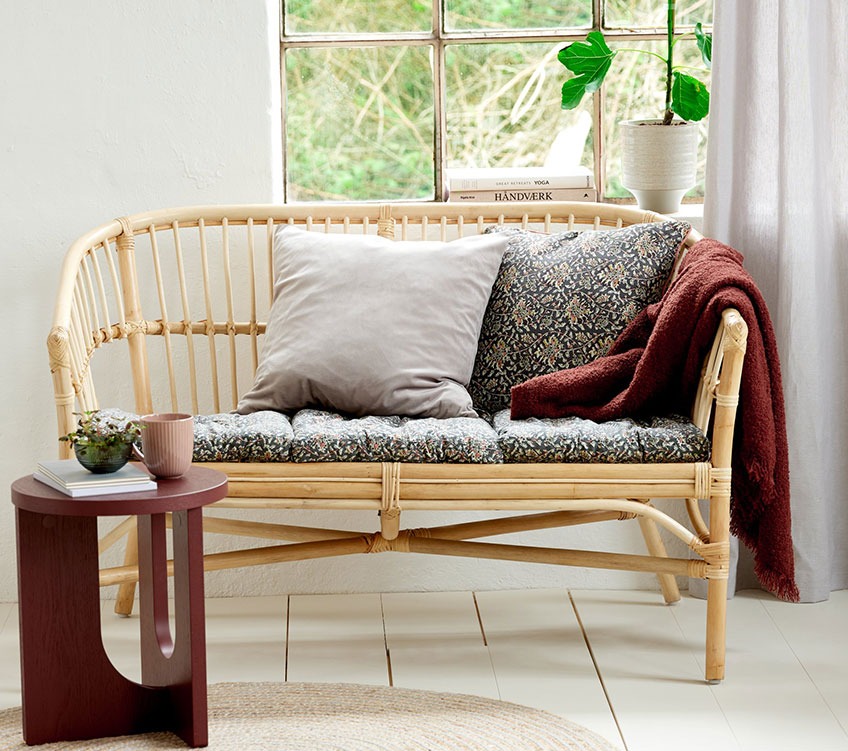 Διθέσιος καναπές από ρατάν με μαξιλάρια καθίσματος και διακοσμητικά μαξιλάρια πλάτης, ένα ριχτάρι σε χρώμα βουργουνδίας και ένα στρογγυλό βοηθητικό τραπεζάκι