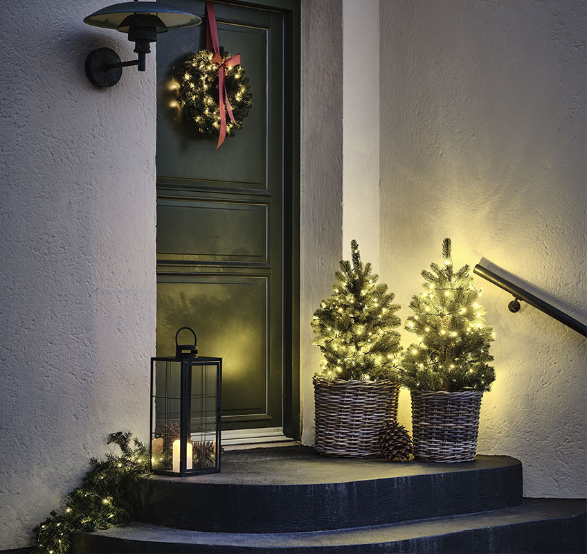 Δυο χριστουγεννιάτικα δεντράκια με λαμπάκια μπροστά σε είσοδο σπιτιού και χριστουγεννιάτικο στεφάνι με κόκκινο φιόγκο